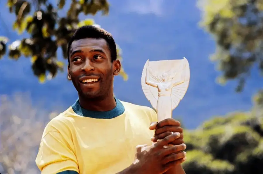 A smiling man in a yellow shirt holds a trophy shaped like a winged figure. He is outdoors, with trees and a blue sky in the background.