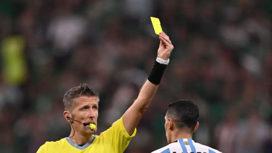 A soccer referee in a yellow shirt shows a yellow card while blowing a whistle. A player in a white and blue striped jersey stands in the foreground with his back to the camera.
