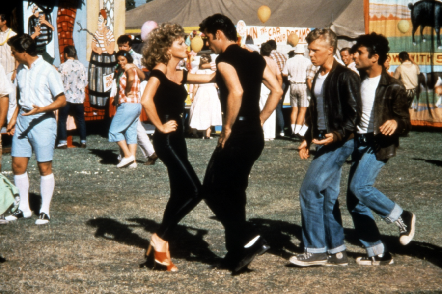 Two people in black outfits dance together at an outdoor fair, with colorful tents and other casually dressed people in the background. The scene is lively and festive.