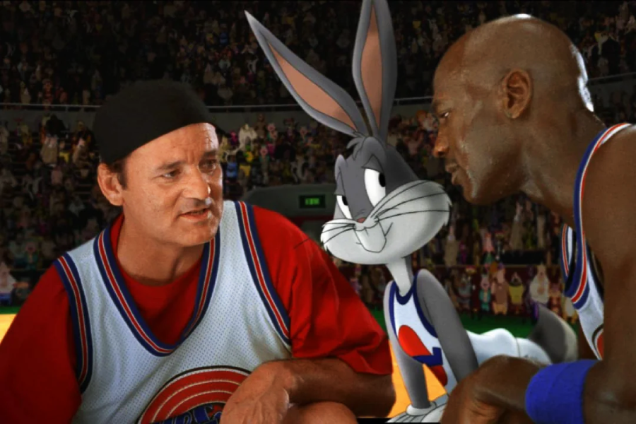 A man in a red shirt and white basketball jersey talks to another man in a jersey, with Bugs Bunny standing between them, during a crowded basketball game.