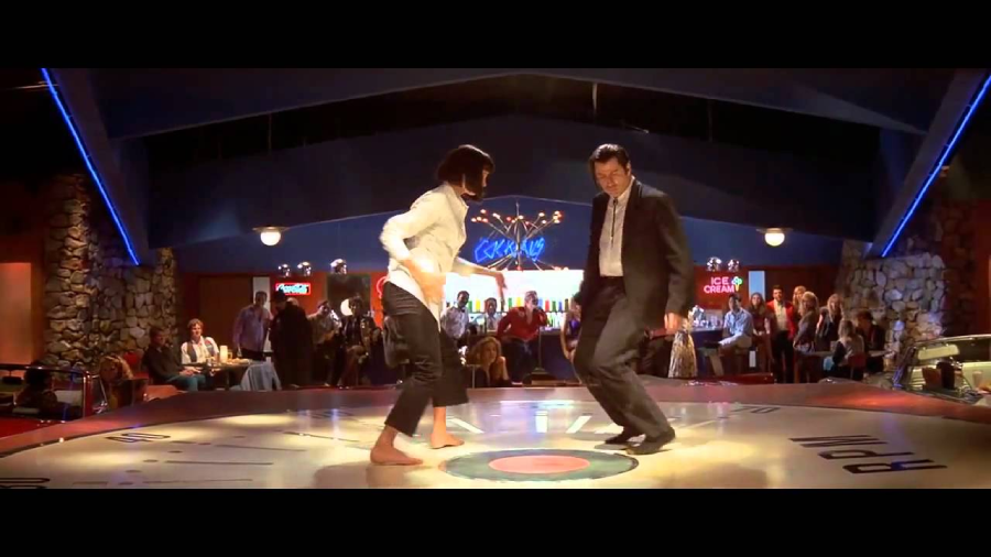 A woman and a man dance together on a stage in a retro-style diner, surrounded by an audience. The man wears a suit and the woman is barefoot with cropped pants and a white shirt. The atmosphere is lively and colorful.