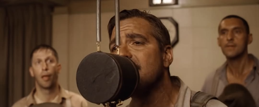 Three men sing into a vintage microphone in a recording studio, with the man in the center in focus and two others in the background. The setting suggests an old-fashioned or historical time period.