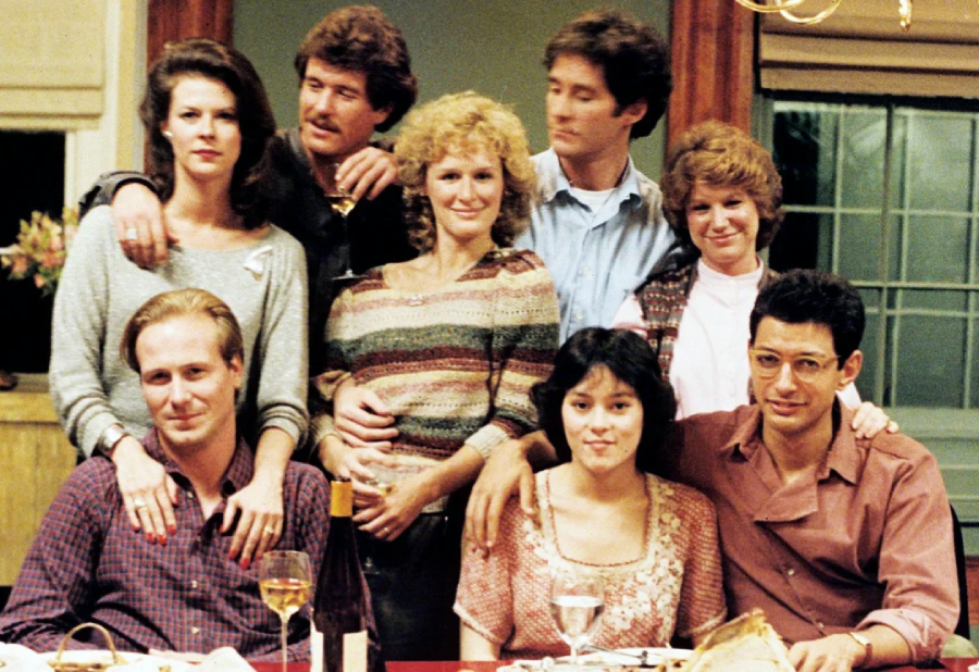 Eight adults pose closely together indoors around a dinner table with wine and food, smiling warmly. The group appears relaxed and friendly, with some people sitting and others standing behind them.