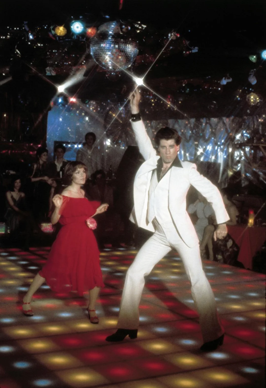 A man in a white suit strikes a dramatic disco pose on a colorful, illuminated dance floor, while a woman in a red dress dances nearby. People watch from tables in a dimly lit club with a mirror ball above.