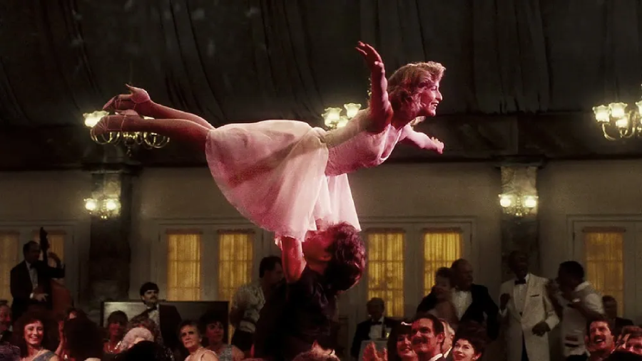 A woman in a white dress is lifted high above a man's head during a dance performance, surrounded by an audience in formal attire watching and applauding.