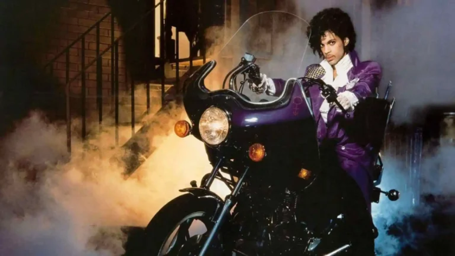 A person in a purple suit sits on a black motorcycle, surrounded by smoke, with stairs and a brick wall in the background.