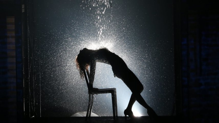 A person leans back dramatically on a chair, silhouetted against bright backlight, as water pours down, creating a striking scene with droplets illuminated in the air.