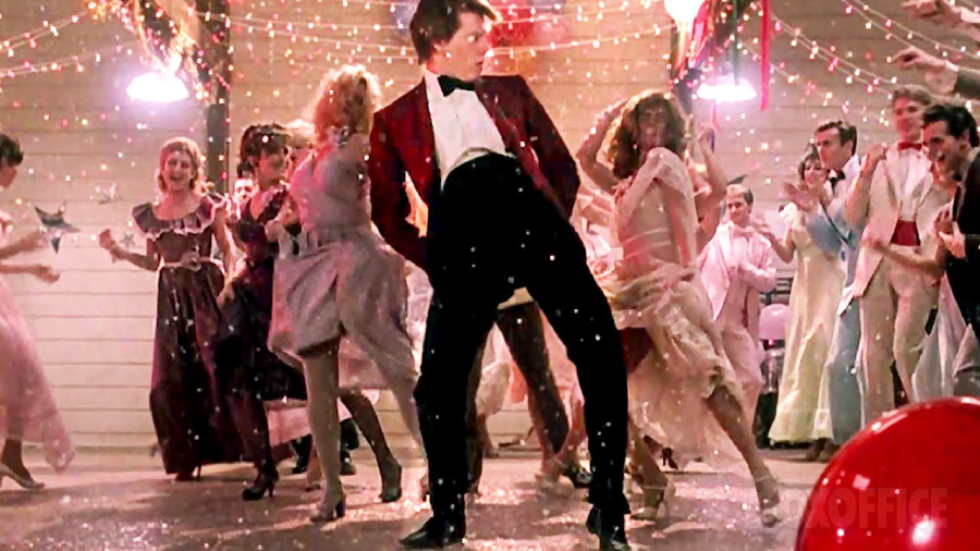 A young man in a red jacket dances energetically at a lively prom, surrounded by people in formal 1980s attire, with string lights and decorations overhead and confetti falling around them.
