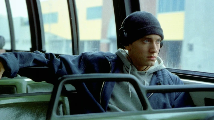 A young man wearing a beanie and headphones sits alone on a bus, looking thoughtfully out the window. He wears a blue jacket and a gray hoodie, with his arm resting on the seat beside him.