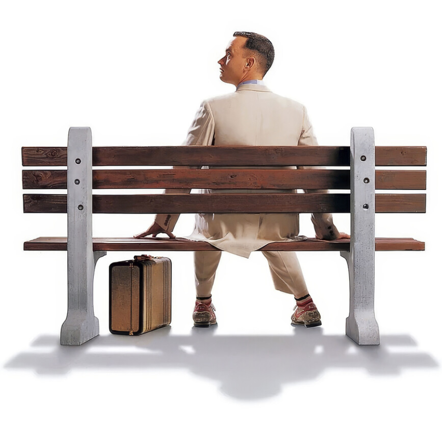 A man in a light suit sits on a wooden bench, viewed from behind, with a suitcase placed on the ground beside him against a plain white background.