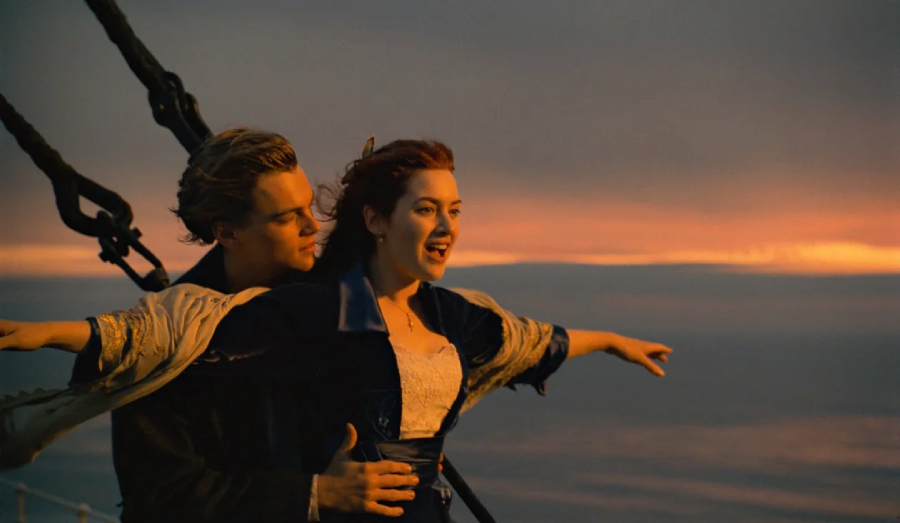 A young couple stands at the bow of a ship at sunset. The woman spreads her arms wide, smiling, as the man stands behind her, holding her gently. The sky is orange and darkening over the ocean.