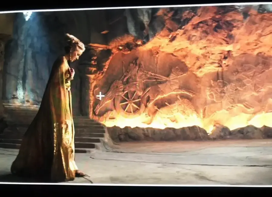 A woman in a gold robe stands in profile, gazing at a large, glowing stone wall engraved with ancient symbols and a wheel, in a dimly lit, cavernous room.