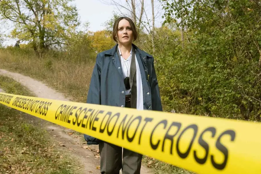 A woman wearing a blue coat stands behind yellow "Crime Scene Do Not Cross" tape on a dirt path surrounded by trees and greenery in an outdoor setting.