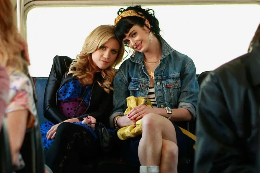 Two young women sit closely together on a bus seat, smiling. One has blonde hair and wears a blue dress, while the other has dark hair, a headband, and a denim jacket. They look relaxed and happy.