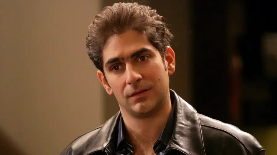 A man with short, dark, wavy hair is wearing a black leather jacket and looking slightly to the side with a neutral expression, standing indoors against a blurred background.