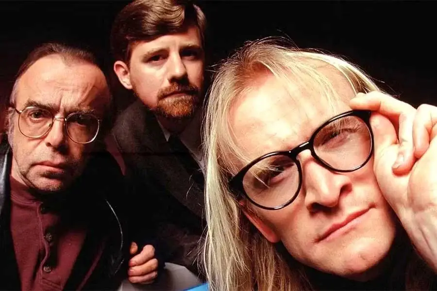 Three men pose closely together, all wearing glasses. The man in front has long blond hair and is adjusting his glasses, while the two men behind have short hair and serious expressions.