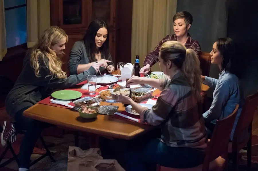 Five women sit around a dining table sharing a meal, talking and smiling. Food containers and drinks are spread across the table, creating a warm, casual, and friendly atmosphere.