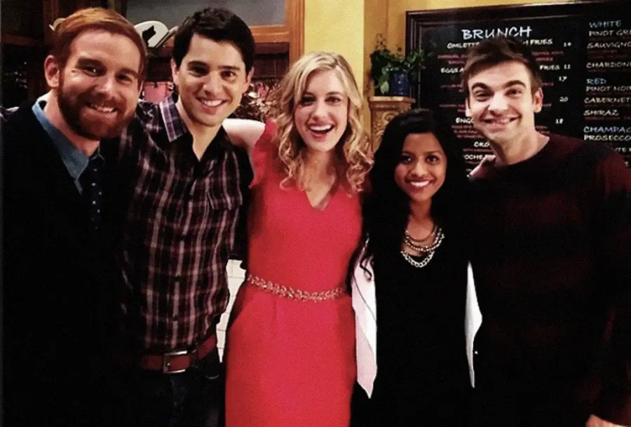 Five young adults smiling and posing together indoors; three men and two women stand in a line with arms around each other, with a chalkboard brunch menu in the background.