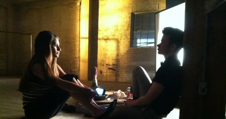 Two people sit facing each other on a wooden floor in a dimly lit room with exposed brick walls and a large window, talking. A bottle, takeout food, and a phone are placed between them.