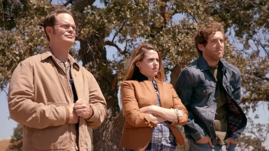 Three people stand outdoors in front of a large tree. The man on the left wears glasses and a tan jacket, the woman in the center has long hair and folds her arms, and the man on the right wears a denim jacket.