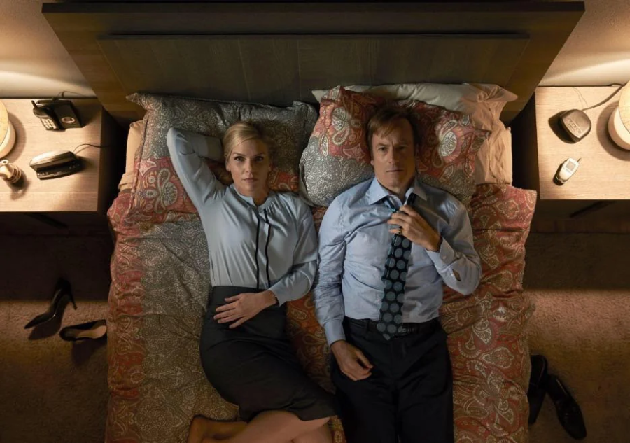 A woman and a man in business attire lie side by side on a bed, staring up at the ceiling. Their shoes are on the floor beside the bed, and the room looks tidy, with matching lamps and nightstands on each side.