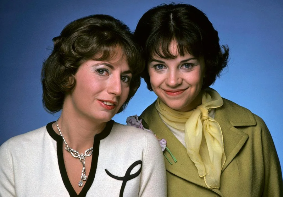 Two women with brown hair pose together, smiling at the camera. One wears a white sweater with a black trim and an "L" on it; the other wears a light green coat and yellow scarf. The background is blue.
