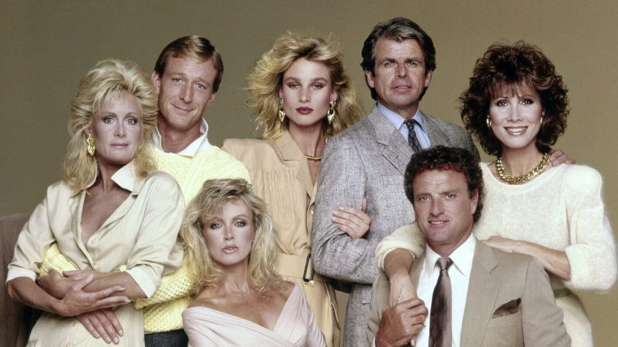 A group of seven adults, dressed in light-colored, stylish 1980s clothing, pose together in front of a plain background, smiling and looking at the camera.