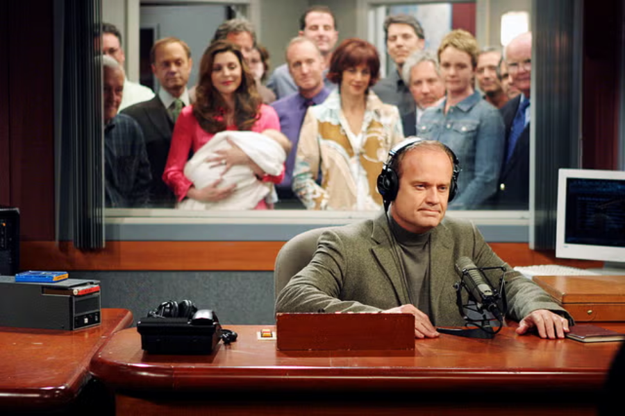 A man wearing headphones sits at a radio studio desk, speaking into a microphone. Behind a glass window, a group of people, including a woman holding a baby, watch him from the adjacent room.