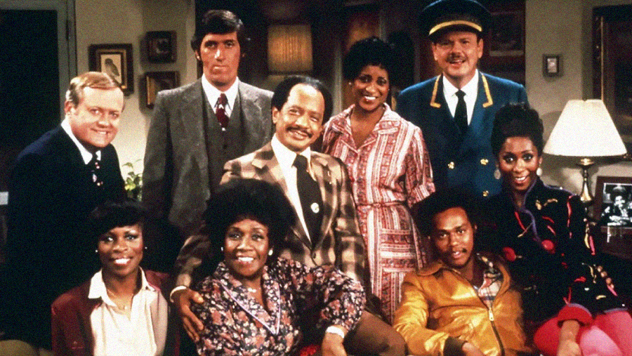 A group of nine adults pose together in a living room setting, dressed in 1970s-style clothing. They are smiling and grouped closely, with a lamp and framed photos in the background.