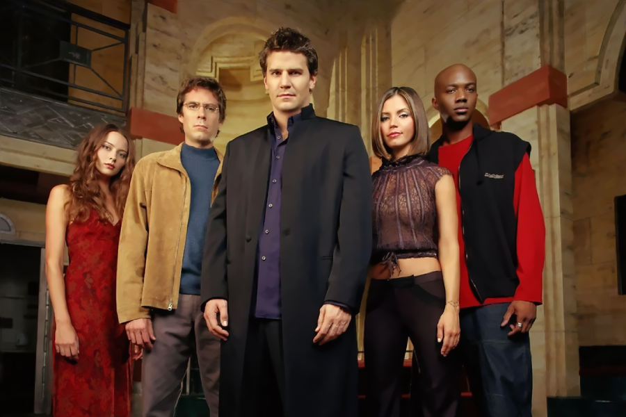 Five people stand together indoors, looking serious. The central man wears a long dark coat; others wear casual or stylish outfits. The background features marble walls and a staircase, suggesting an elegant, dramatic setting.
