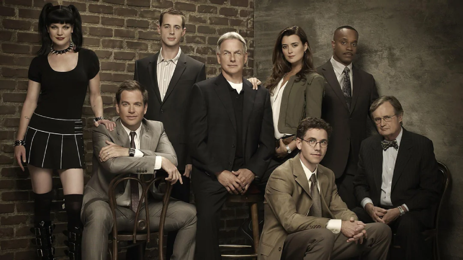 Eight people pose together in front of a brown brick wall, dressed in professional and casual attire, with serious expressions, suggesting they are part of a team or cast ensemble.