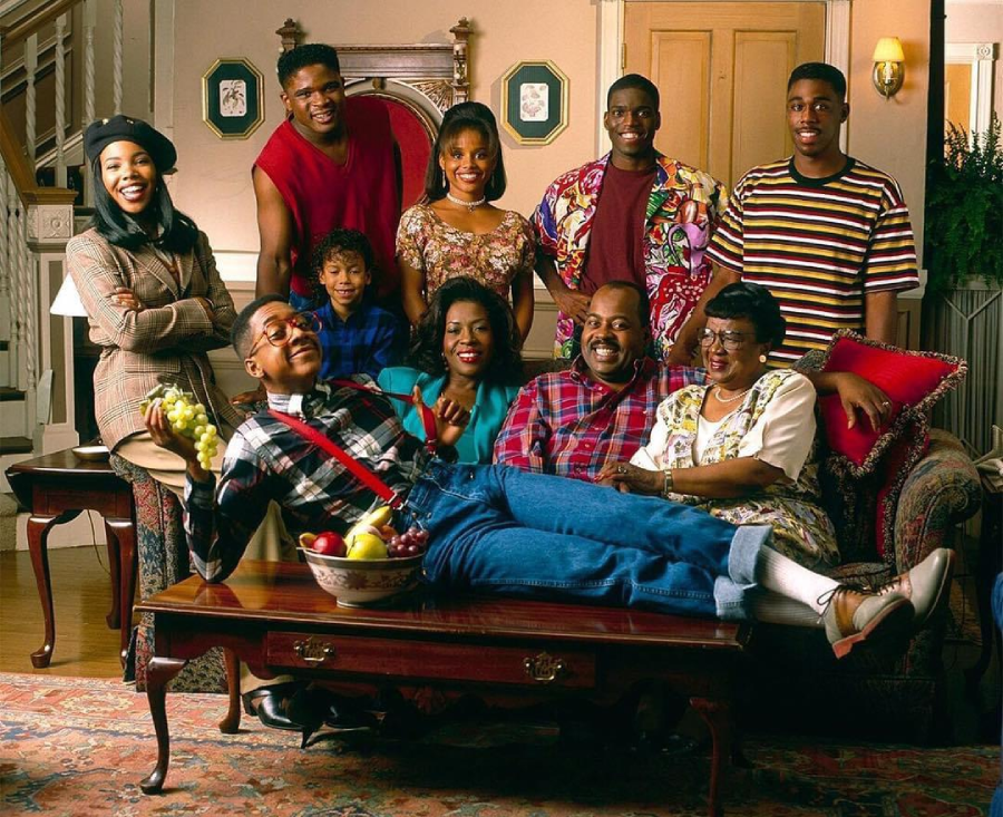 A group of nine people pose and smile in a cozy living room. One person in suspenders lies playfully across a coffee table while the rest sit or stand around him, creating a joyful and lively atmosphere.