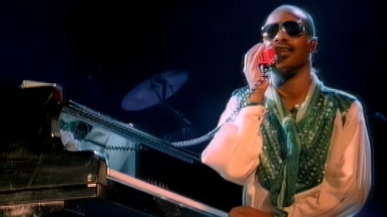 A musician wearing sunglasses and a green vest sings into a red microphone while playing a piano on a dimly lit stage.