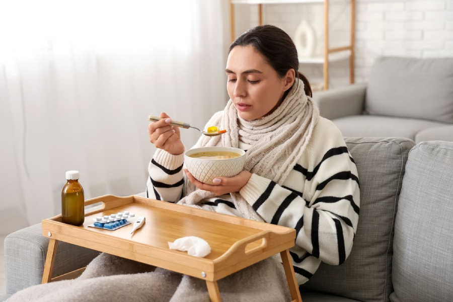 A woman wrapped in a scarf sits on a couch with a blanket, eating soup from a bowl. A wooden tray on her lap holds medicine, tissues, and a bottle, suggesting she is unwell and resting at home.