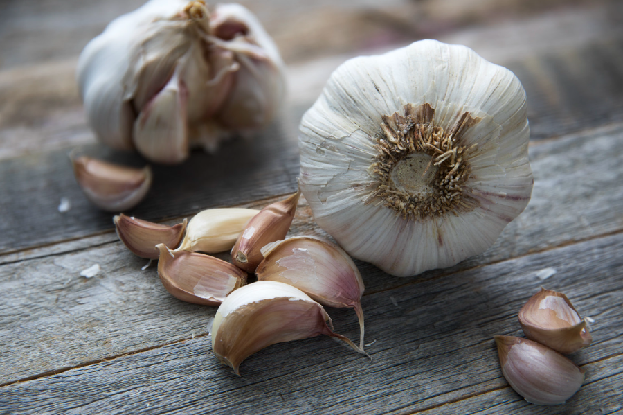 Two heads of garlic and several individual garlic cloves are scattered on a rustic wooden surface. The cloves have light brown papery skins and the scene is lit with natural light.