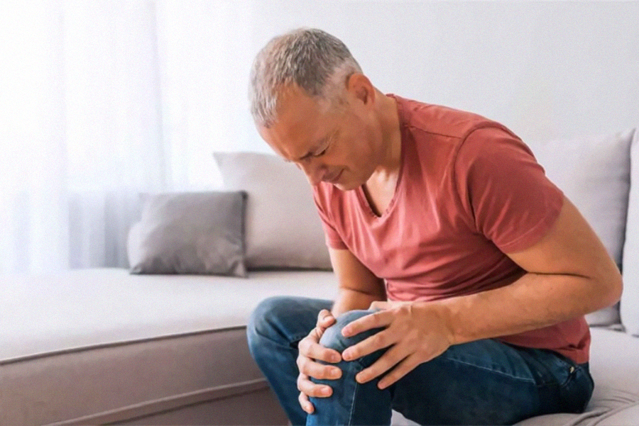 A middle-aged man sits on a couch, holding his knee and wincing in pain, suggesting discomfort or injury. The room is softly lit, with neutral-colored furniture in the background.