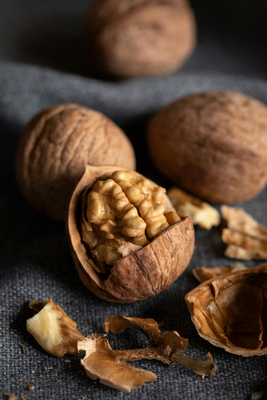 A close-up of walnuts, one partially cracked open to reveal the nut inside, with pieces of shell and whole walnuts in the background on a dark fabric surface.