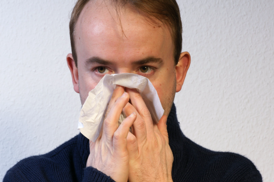 A person in a dark sweater holds a tissue to their nose, appearing to sneeze or blow their nose, against a plain light-colored background.