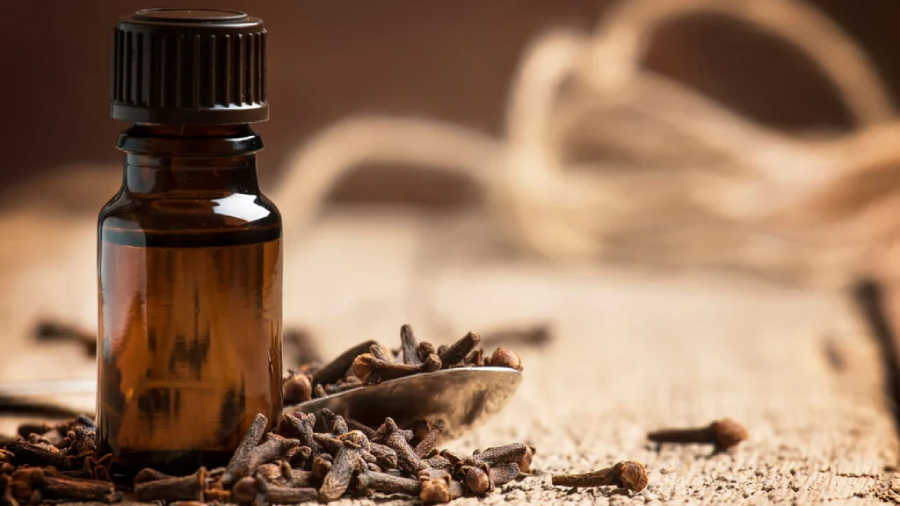 A small amber glass bottle with a black cap sits next to a spoon filled with whole cloves, with more cloves scattered on a wooden surface. A coil of rope is blurred in the background.