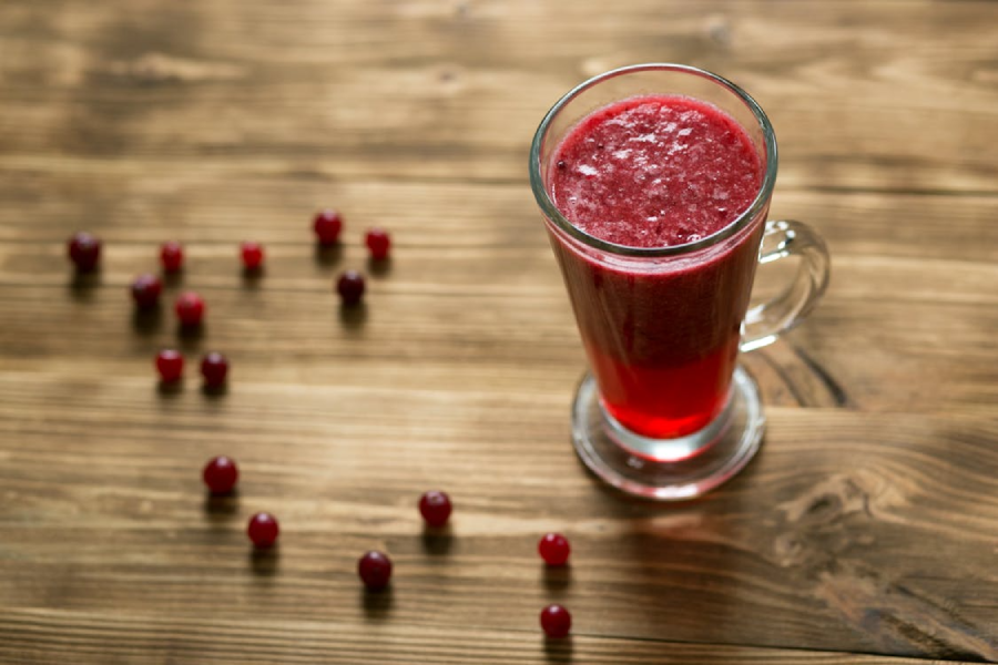 A clear glass mug filled with red berry juice sits on a wooden surface, surrounded by scattered whole red berries. The juice looks thick and freshly blended.
