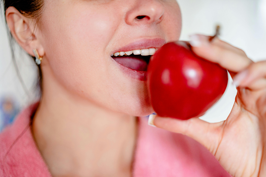 A close-up of a woman wearing a pink robe, about to bite into a shiny red apple. Her mouth is slightly open, showing her teeth, and she is holding the apple near her lips.