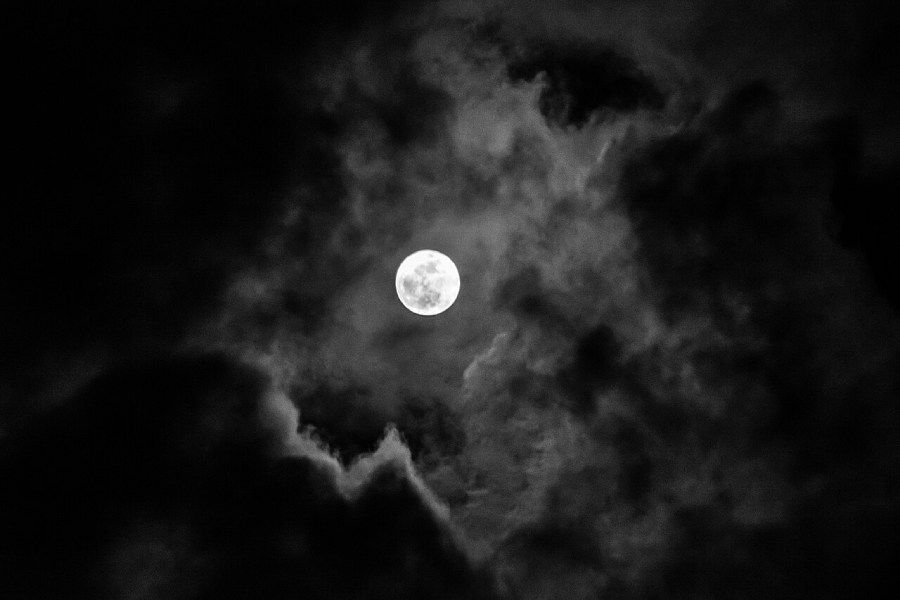 A bright full moon shines in the night sky, surrounded by dark clouds that partially obscure its light.