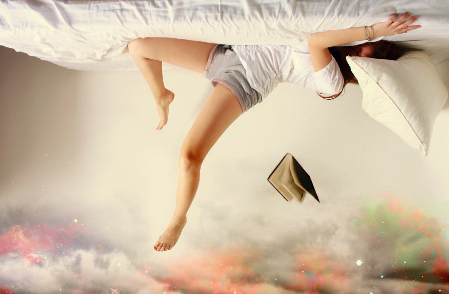 A woman lies upside down on a bed with her legs extending off the edge, a book floating nearby. Below her appears a surreal, colorful, cloud-like scene, blending reality with dreamlike fantasy.