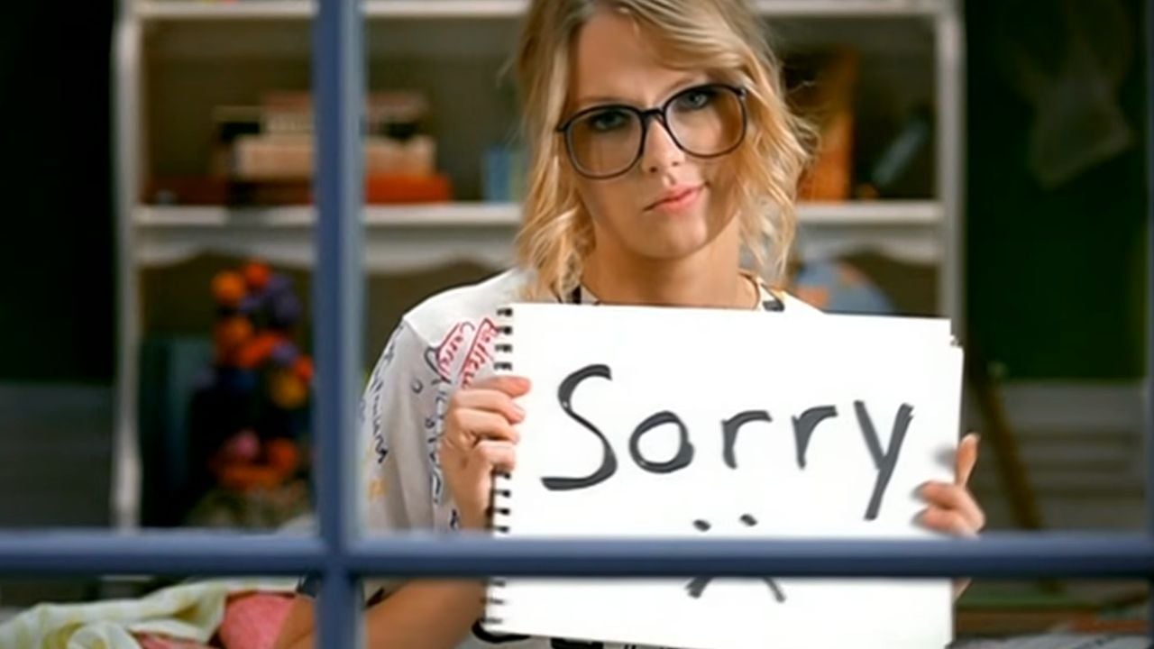 A person with glasses holds a white sign with “Sorry” and a sad face drawn on it, looking through a window with a neutral expression. Shelves with books and decorations are visible in the background.