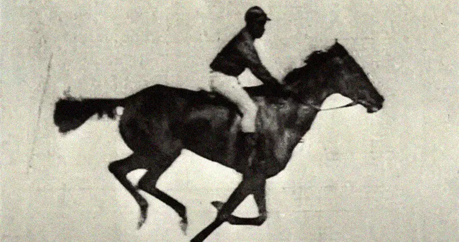 A black-and-white, vintage-style image shows a jockey riding a galloping horse, captured mid-stride with all four of the horse’s legs off the ground. The background is plain and features no visible scenery.