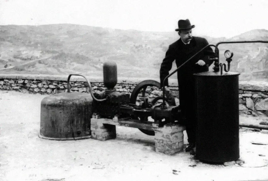 A man in a suit and hat operates early industrial machinery outdoors, with hills in the background. The machine includes large cylinders, pipes, and a gauge on a stone platform.