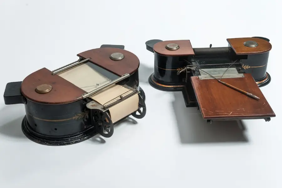 Two antique writing machines with black and copper parts, featuring wooden surfaces, metal rollers, and a pen on one board, set against a plain white background.