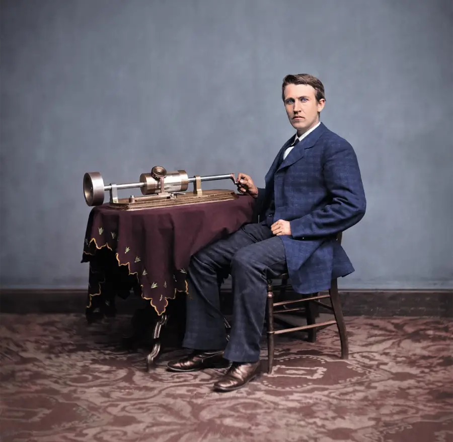 A man in a blue suit sits on a chair beside a table with a vintage mechanical device on it, against a plain blue background and patterned carpet.