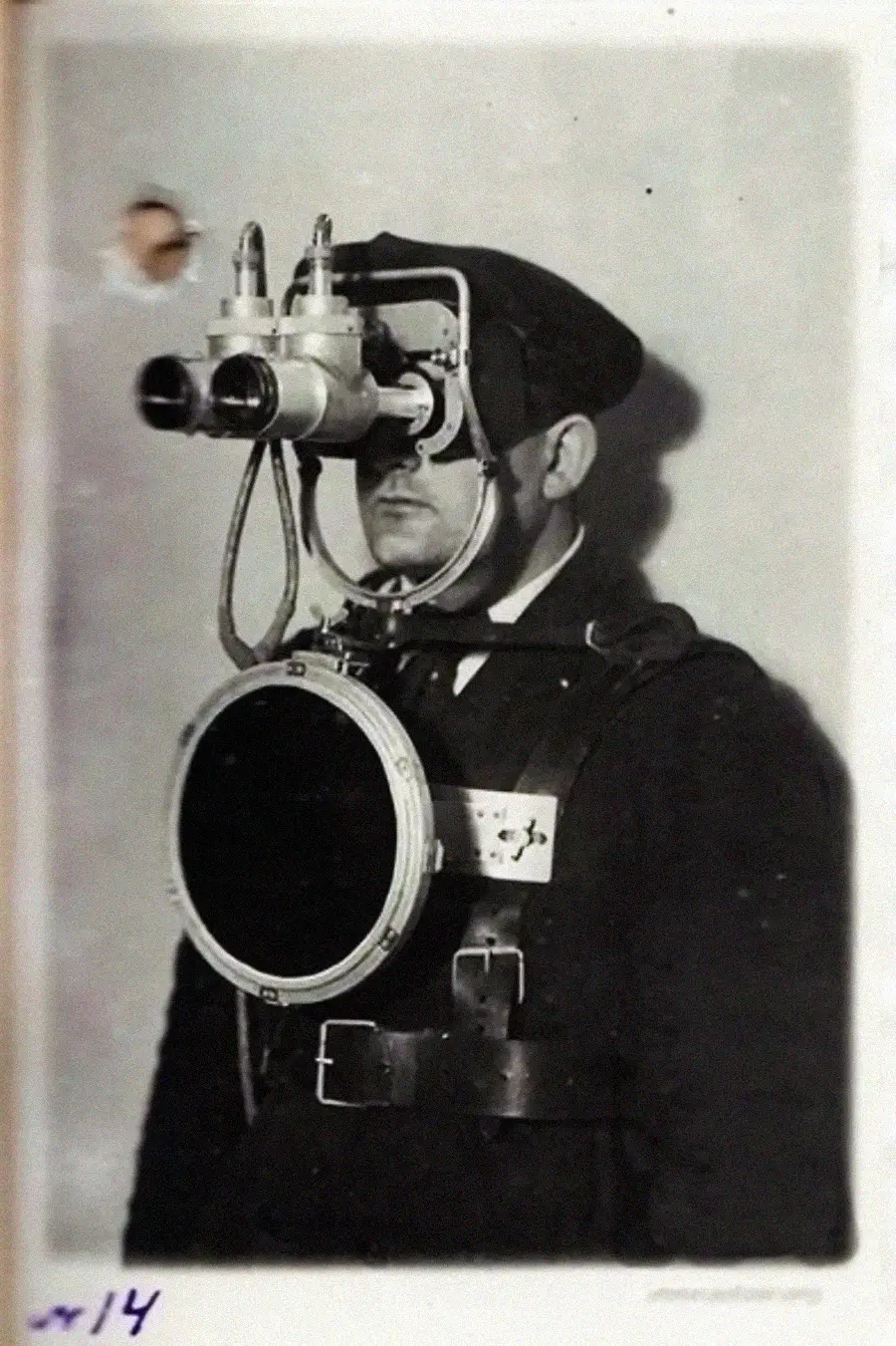 A man in old-fashioned attire wears large binocular goggles attached to his head and a circular device strapped to his chest, suggesting early experimental technology or protective gear.