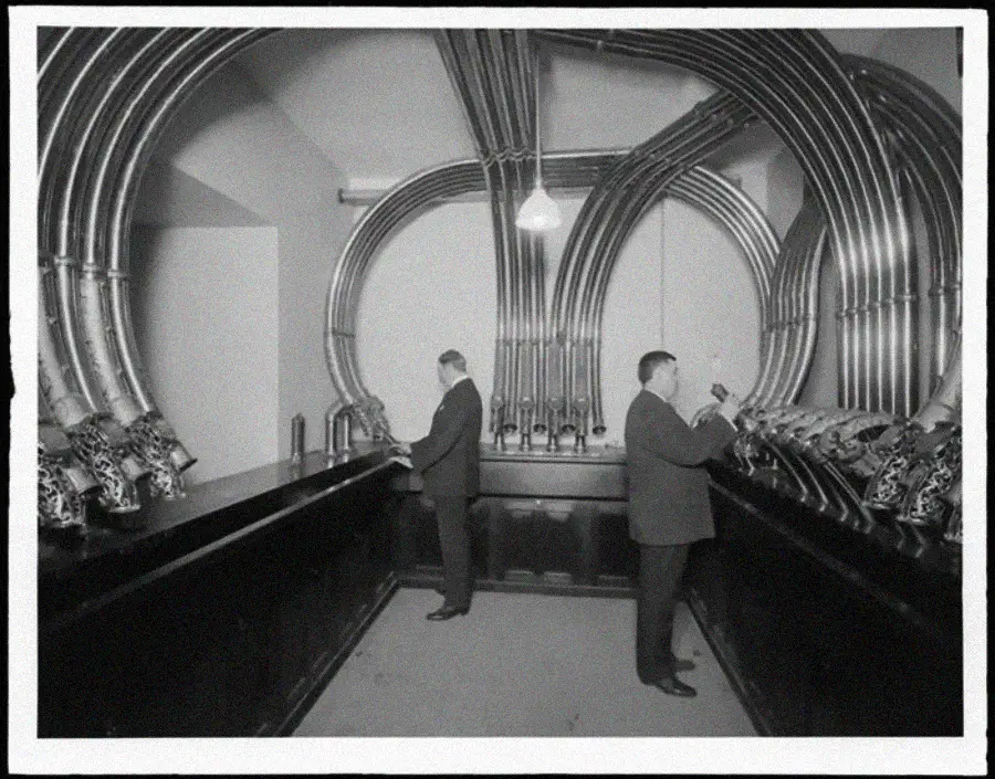 Two men in suits operate control panels in a room with large metal pipes arching along the walls and ceiling, giving the space a futuristic, industrial appearance. Black and white, vintage photo.